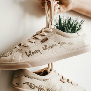 Close-up of beige sneakers with plant design and 'bloom where you are planted' script.